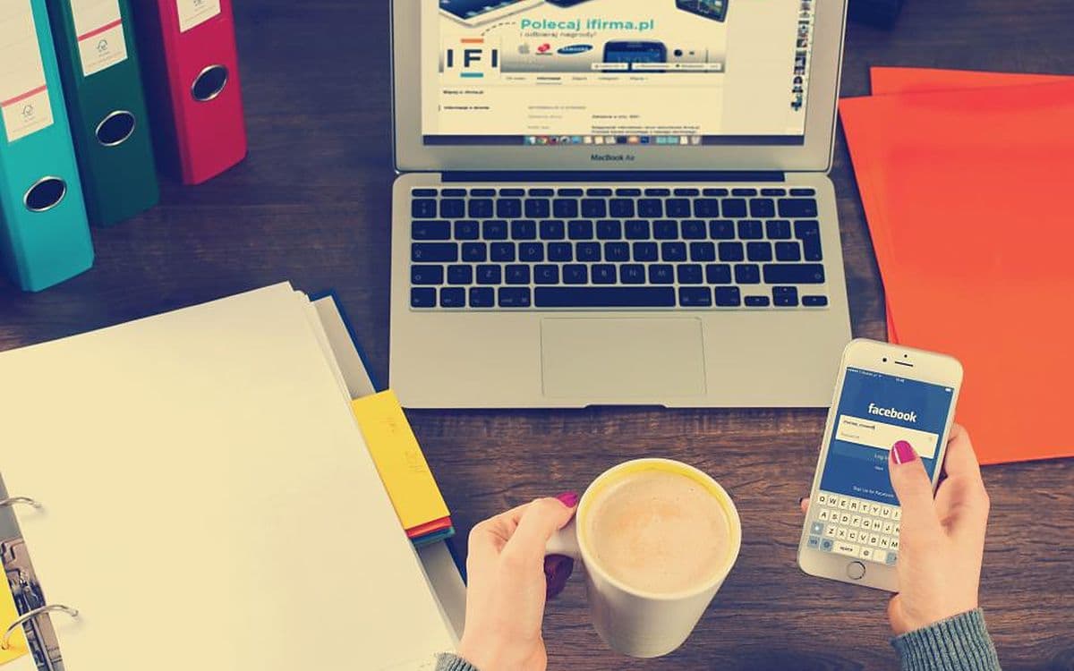 Someone holding a phone open on a social app's sign-in screen, with a laptop, a coffee and colorful folders on the desk.