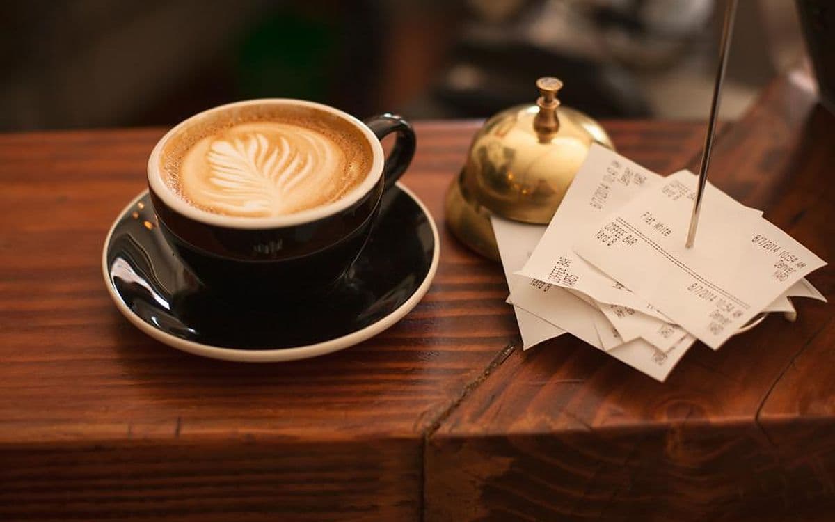 A latte on a café counter next to a brass bell and a spike of paper receipts.