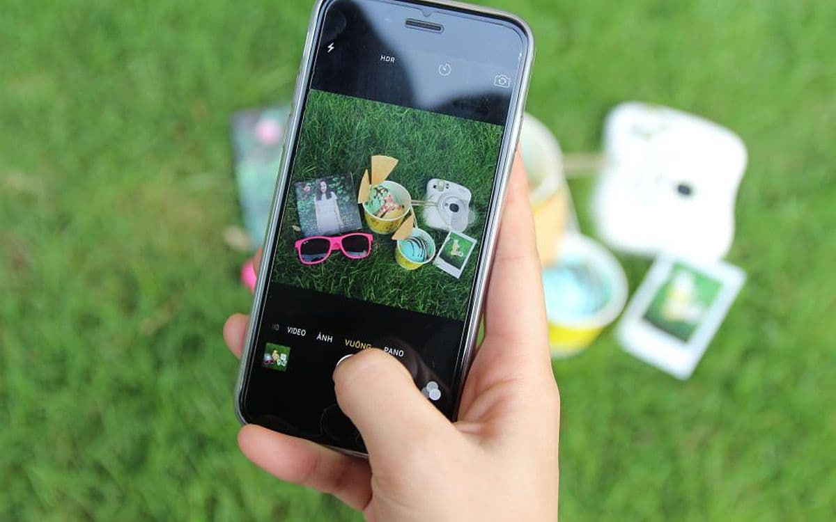 A hand holding a phone over grass scattered with instant photos and sunglasses.