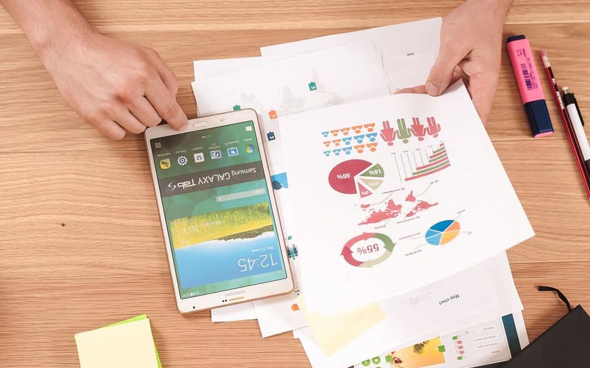 An Android phone on a desk covered in printed charts, two hands working around it.