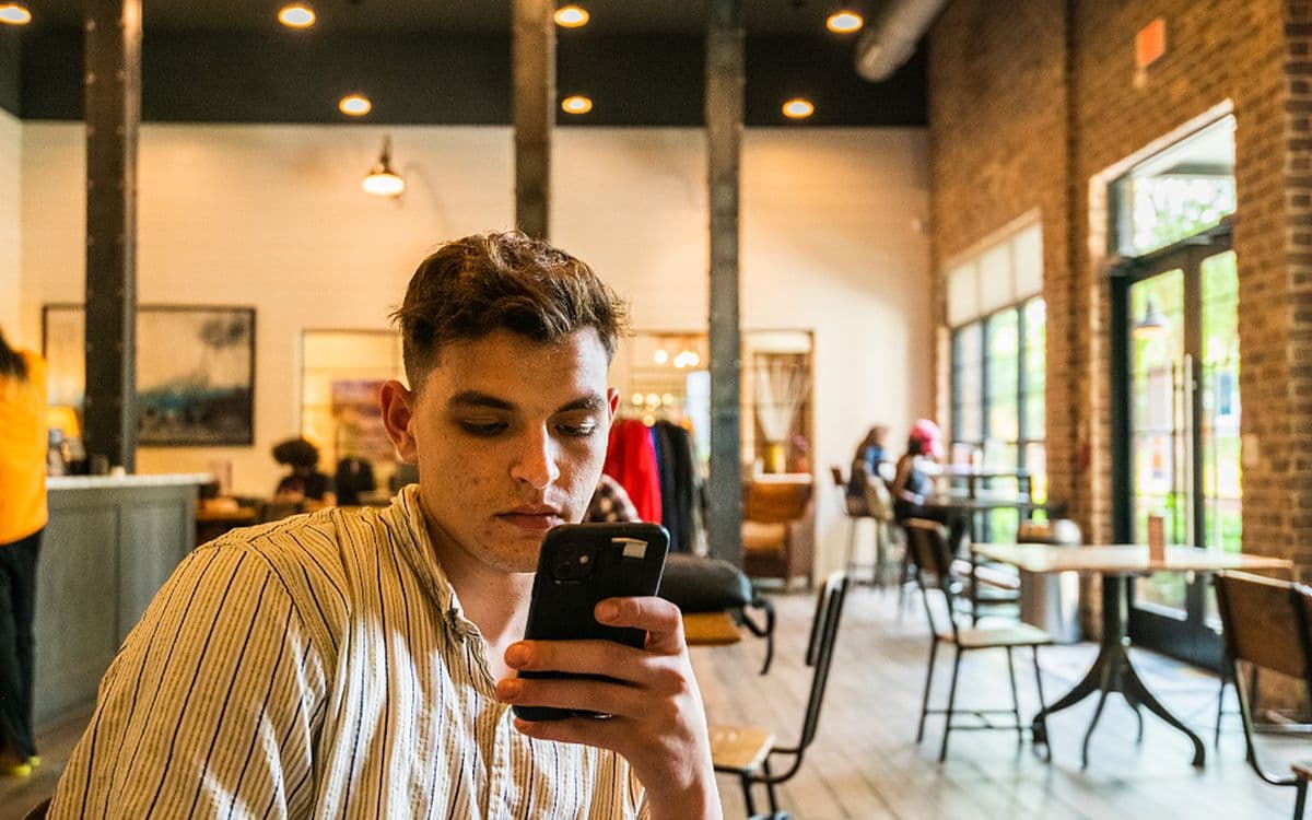 A young man in a busy café looking intently at his phone.