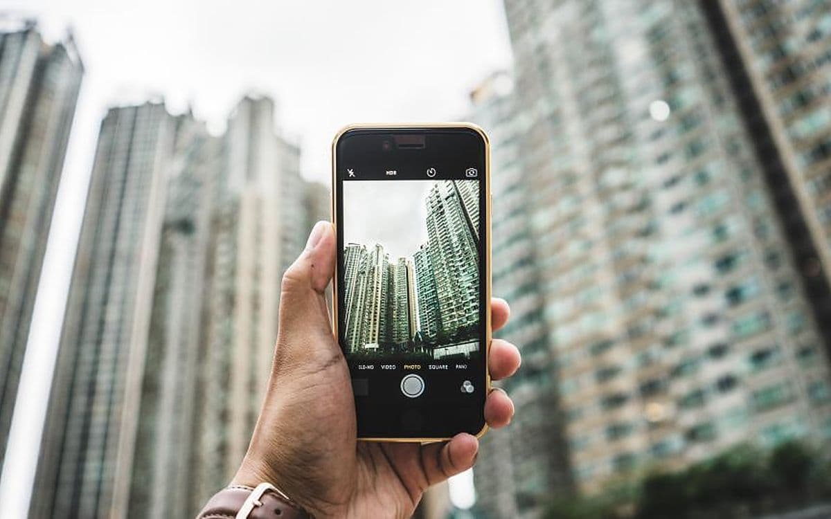 A hand holding a phone up to photograph a row of tall apartment buildings.