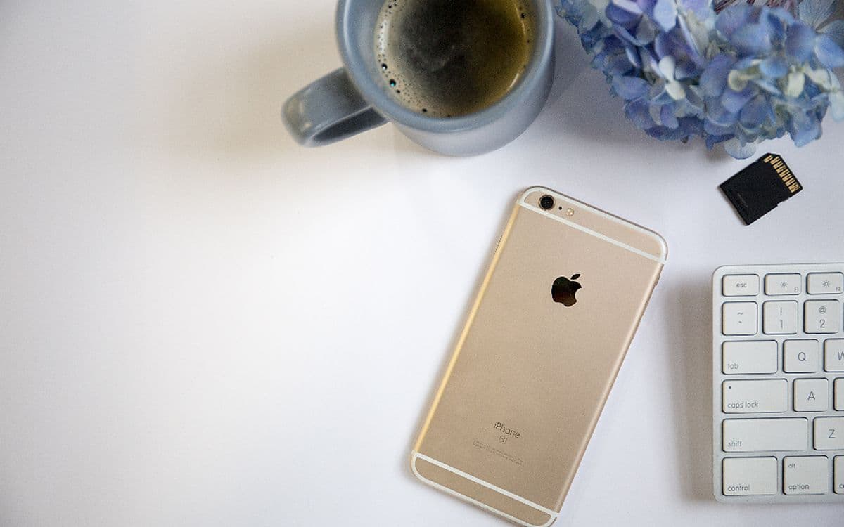 A gold iPhone on a clean white desk beside a mug of coffee, flowers and a keyboard.