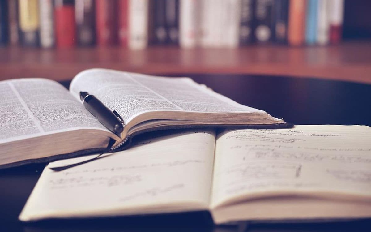 Two open books with handwritten notes and a pen, in front of a shelf of books.