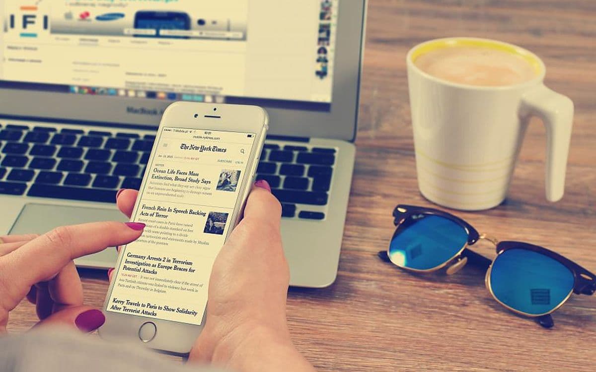 A hand holding an iPhone open on a news article, a laptop, a mug and sunglasses on the desk behind it.