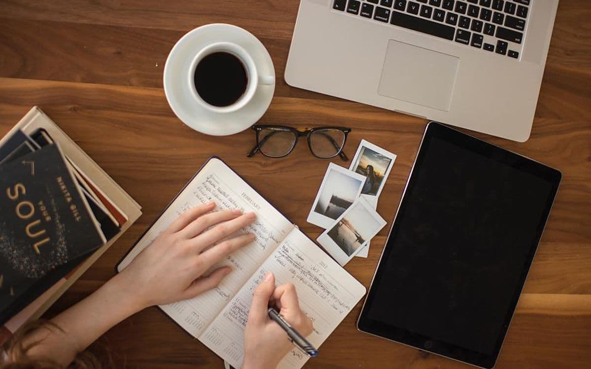 A desk from above with a laptop, a tablet, a notebook, coffee and a few instant photos.