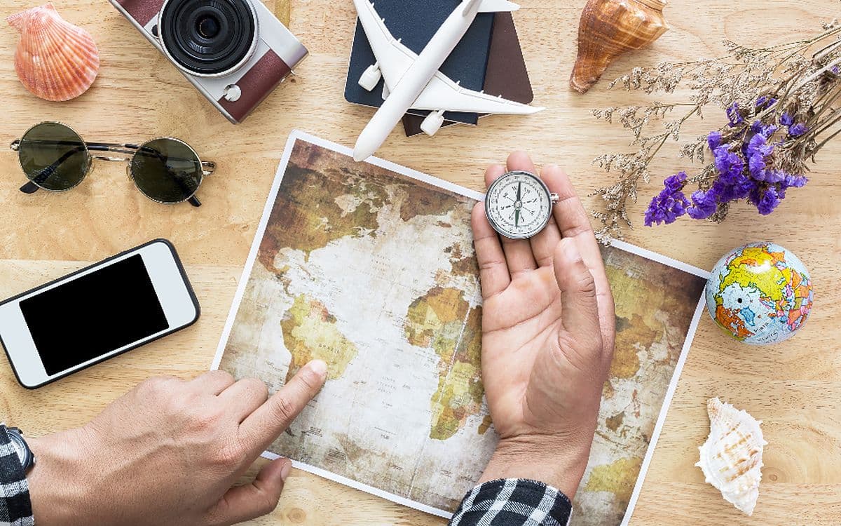 Hands on a world map surrounded by a phone, a camera, sunglasses, a compass and a toy plane.