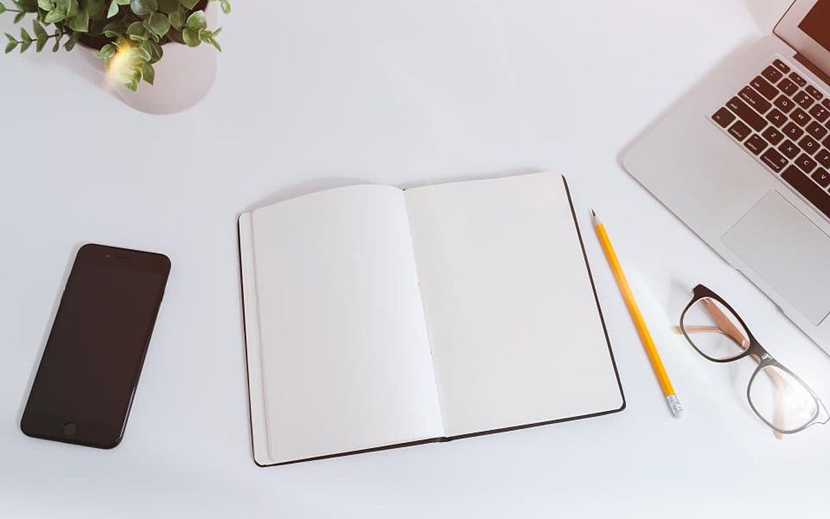 A tidy white desk from above: a phone, an open notebook, a pencil, glasses and a laptop.