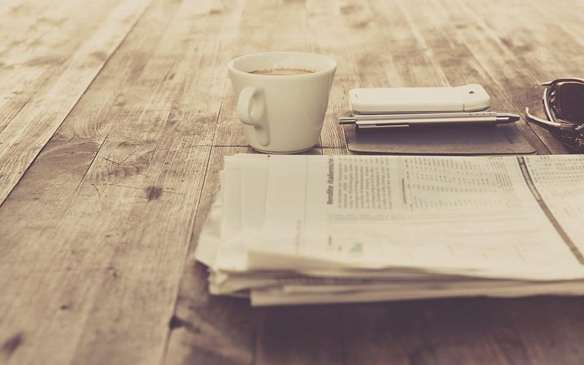 A folded newspaper, a cup of coffee and a phone on a sunlit wooden table.