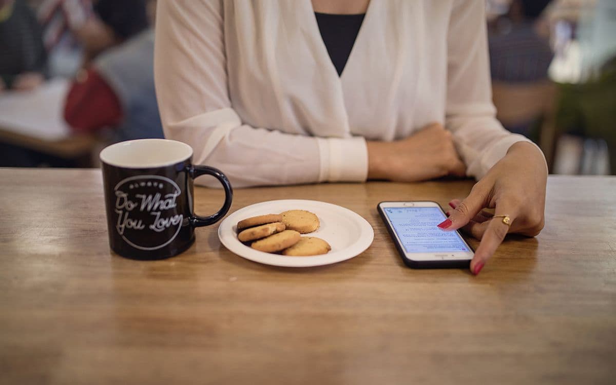 A woman at a café table tapping through something on her phone, a mug and biscuits beside her.